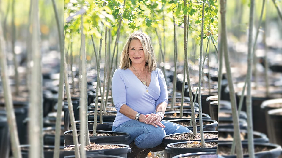 A blonde women kneels in between rows of oak trees growing in black plastic containers.