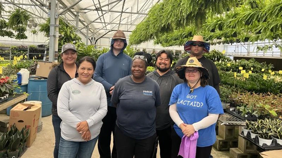 Seven smiling people pose for a photo in a greenhouse surrounded by potted plants and hanging baskets of plants.