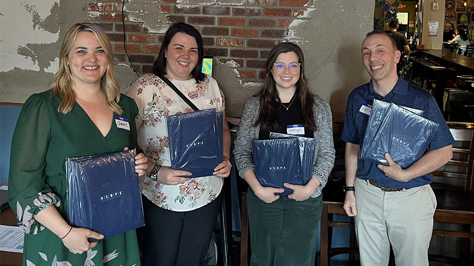 Four smiling people holding blue padded folio awards with an exposed brick wall in background.
