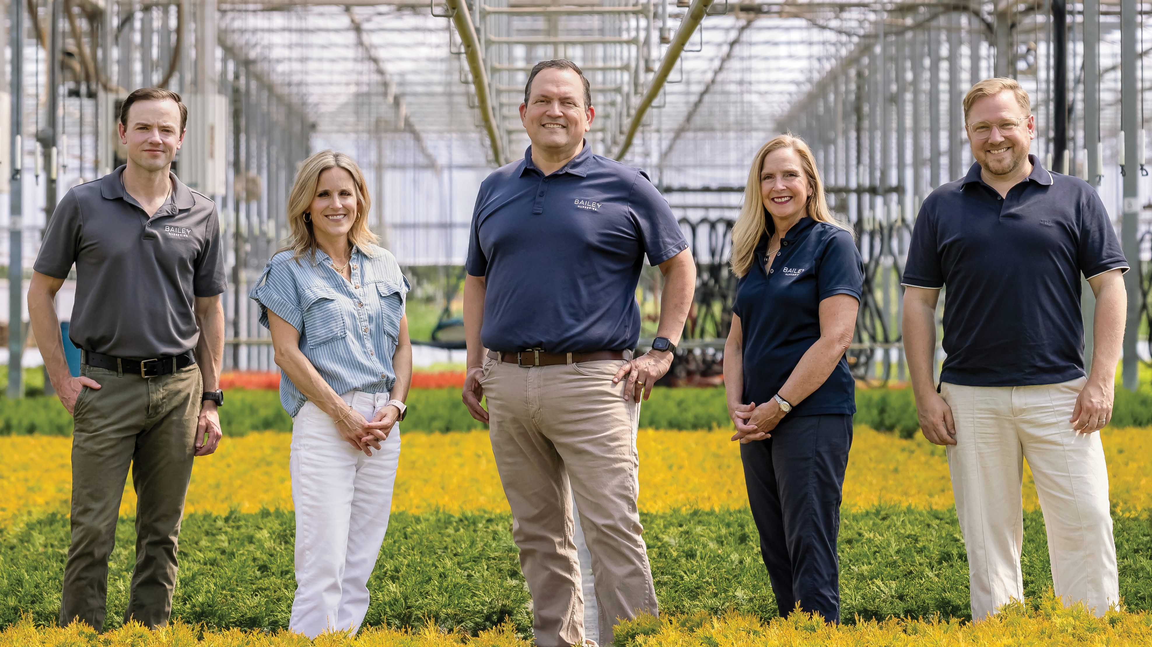 Five smiling people pose in a greenhouse, with yellow and green flowers and plants behind them.