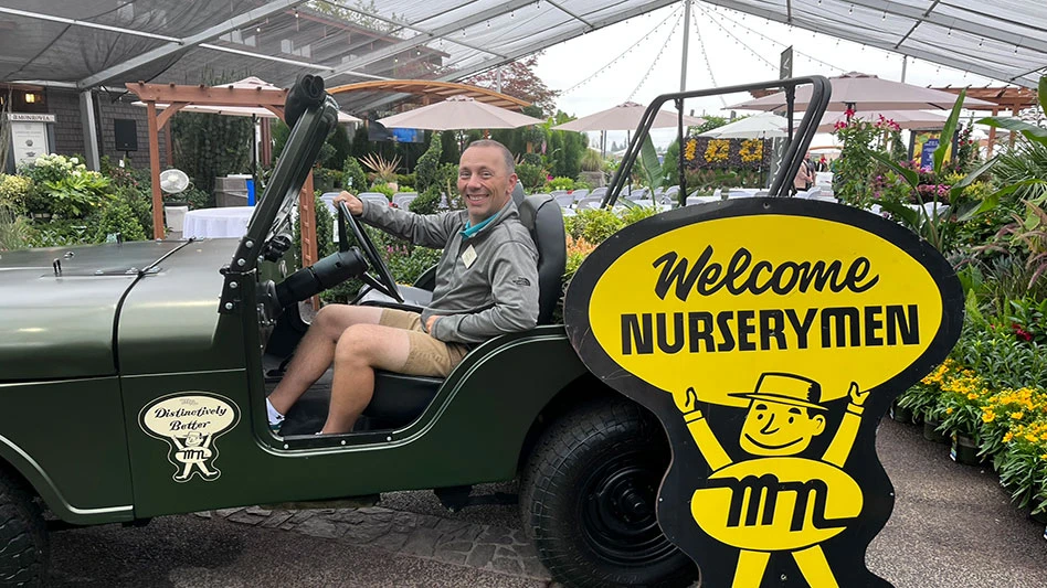 A smiling man sitting in the front seat of a Jeep, which is in front of a yellow and black sign reading Welcome Nurserymen.